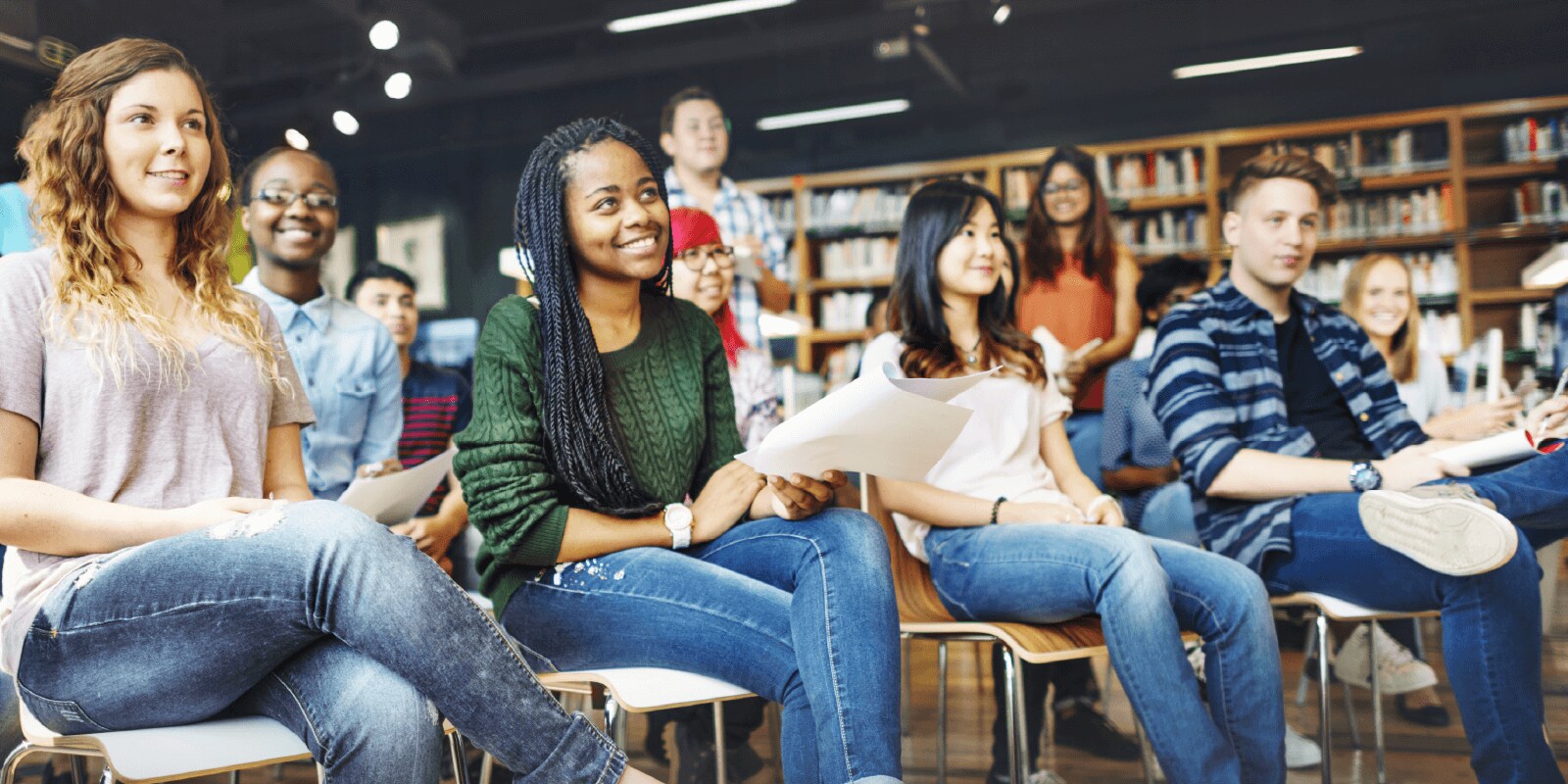 Group of students seated in a modern library classroom, holding papers during an academic discussion