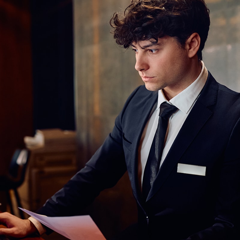 Young man in a suit holding paper looking at a computer screen