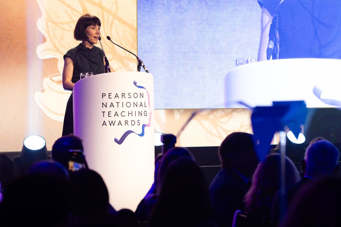 A teacher in classroom of students helping one student with their laptop