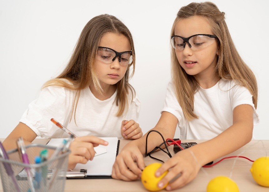 Two pupils sitting at a table carry out a hands-on experiment using lemons connected by wires and a multimeter, with notebooks and pens nearby.