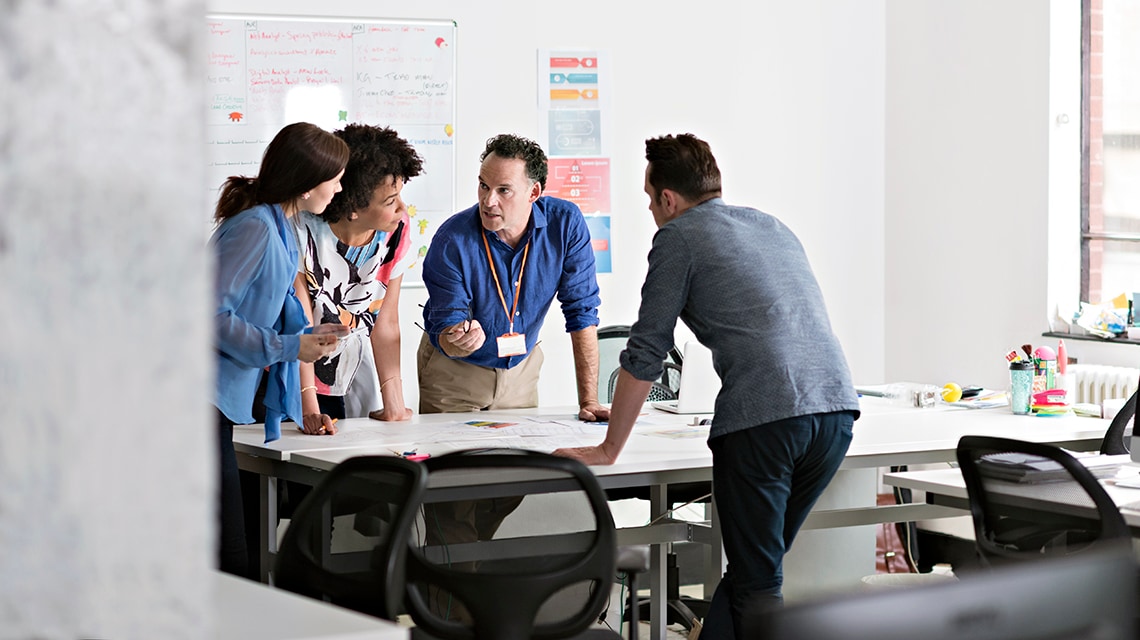 Team of colleagues gathered around a desk in a bright office, reviewing documents and discussing project plans during a collaborative work session.