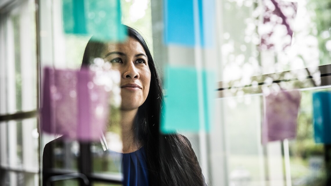 Person analysing colourful sticky notes on a glass wall in a bright office space, reviewing ideas and planning tasks against a backdrop of large windows.