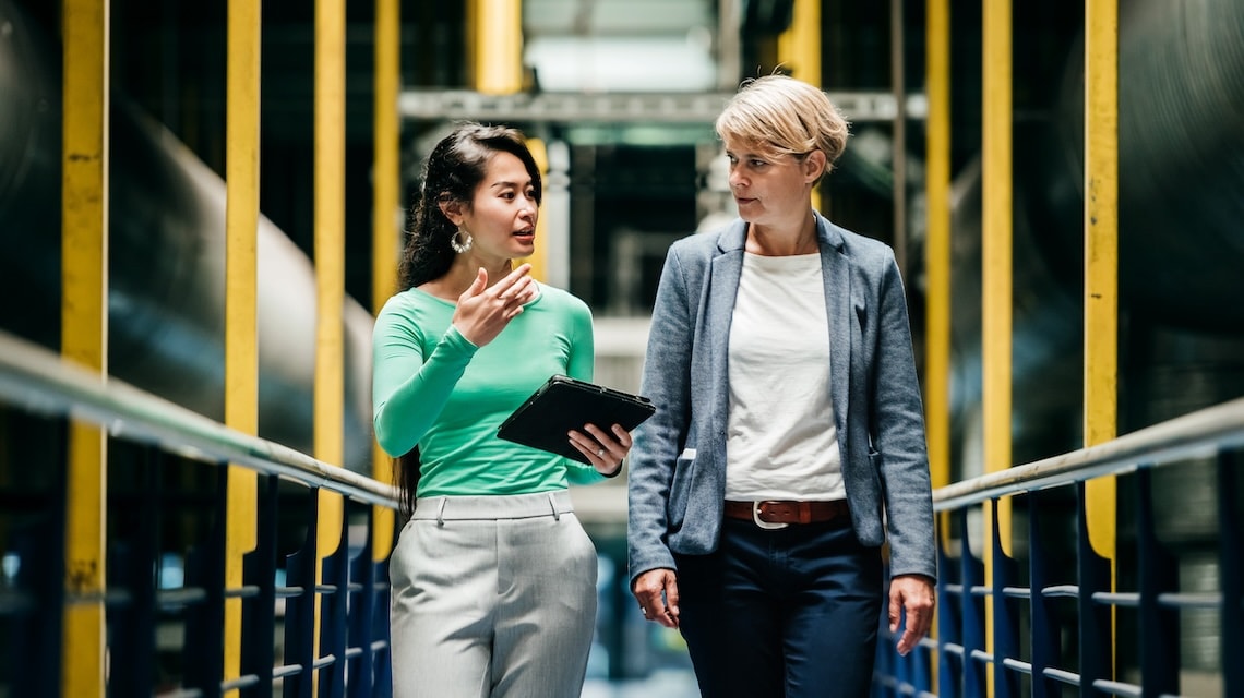 Two colleagues walking through an industrial facility, discussing work while reviewing information on a tablet, surrounded by machinery and yellow safety railings.