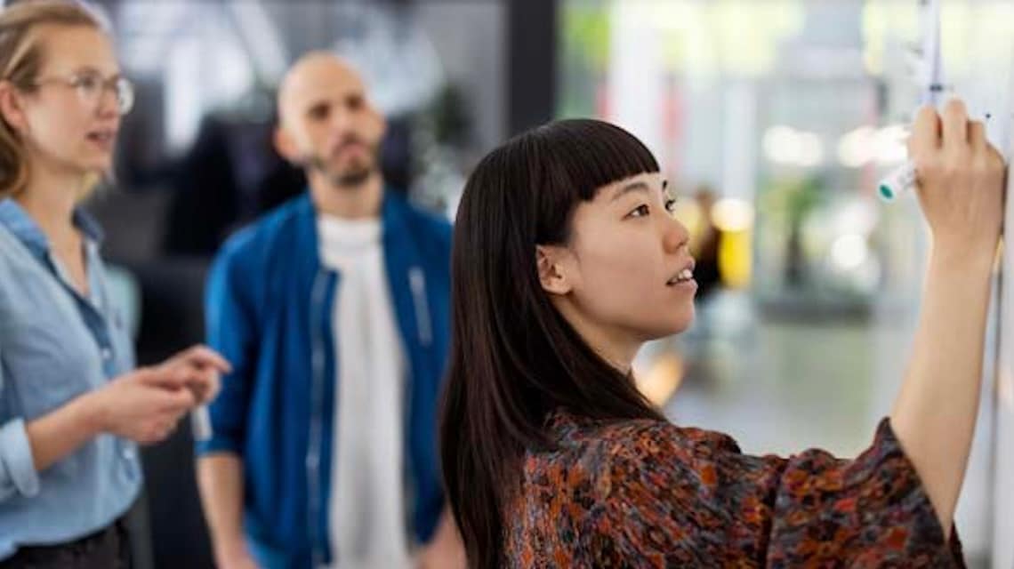 Person writing on a whiteboard during a collaborative session, while colleagues stand nearby discussing ideas in a bright, modern workspace.