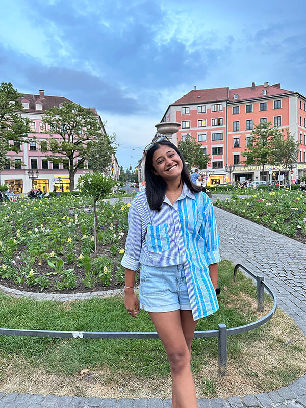 Blog author Arya is outside in her study abroad location, standing in front of a garden with red brick buildings behind it.