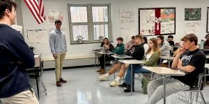 Two male college students standing at the front of a classroom presenting to a group of students seated at desks.