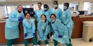 A group of eight nursing students standing in 2 rows. They are all wearing blue scrubs. 