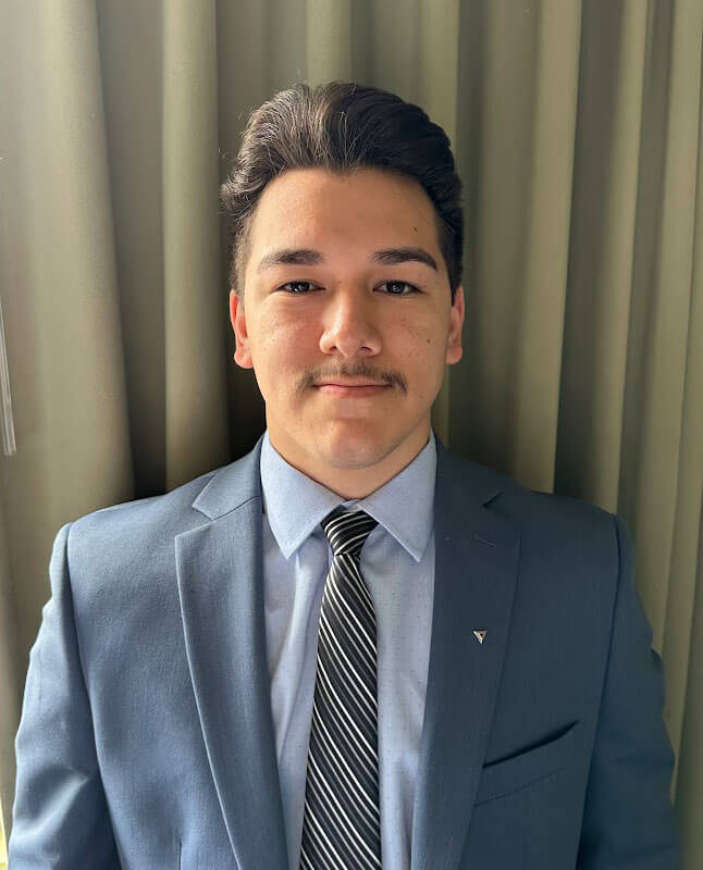 Blog author Aj is standing in front of light green drapes. He has short dark hair and is wearing a grey suit, light blue shirt, and striped tie.