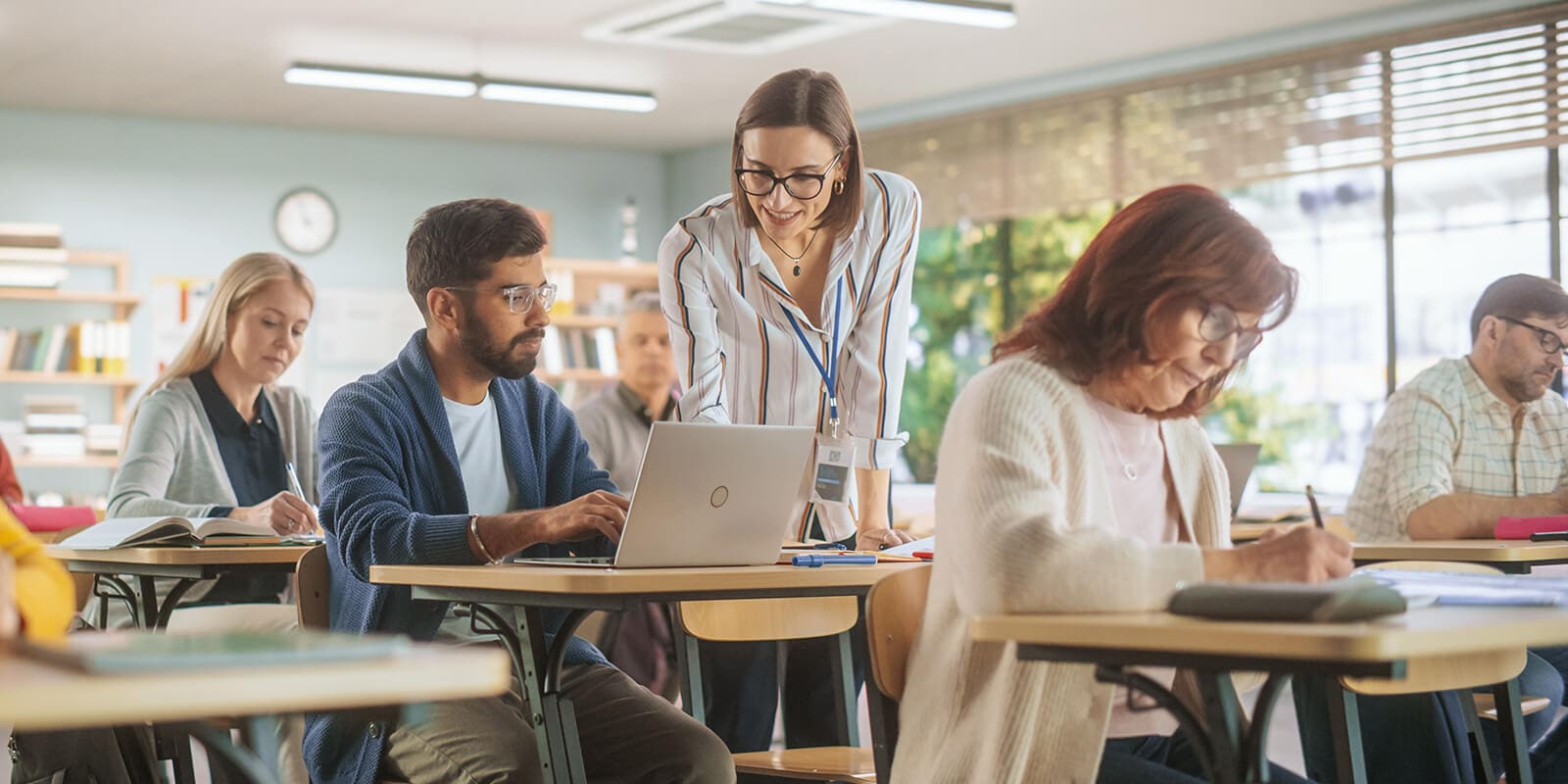Adult students of diverse ages engaged in studying in a brightly lit classroom, some using laptops and others writing in notebooks.