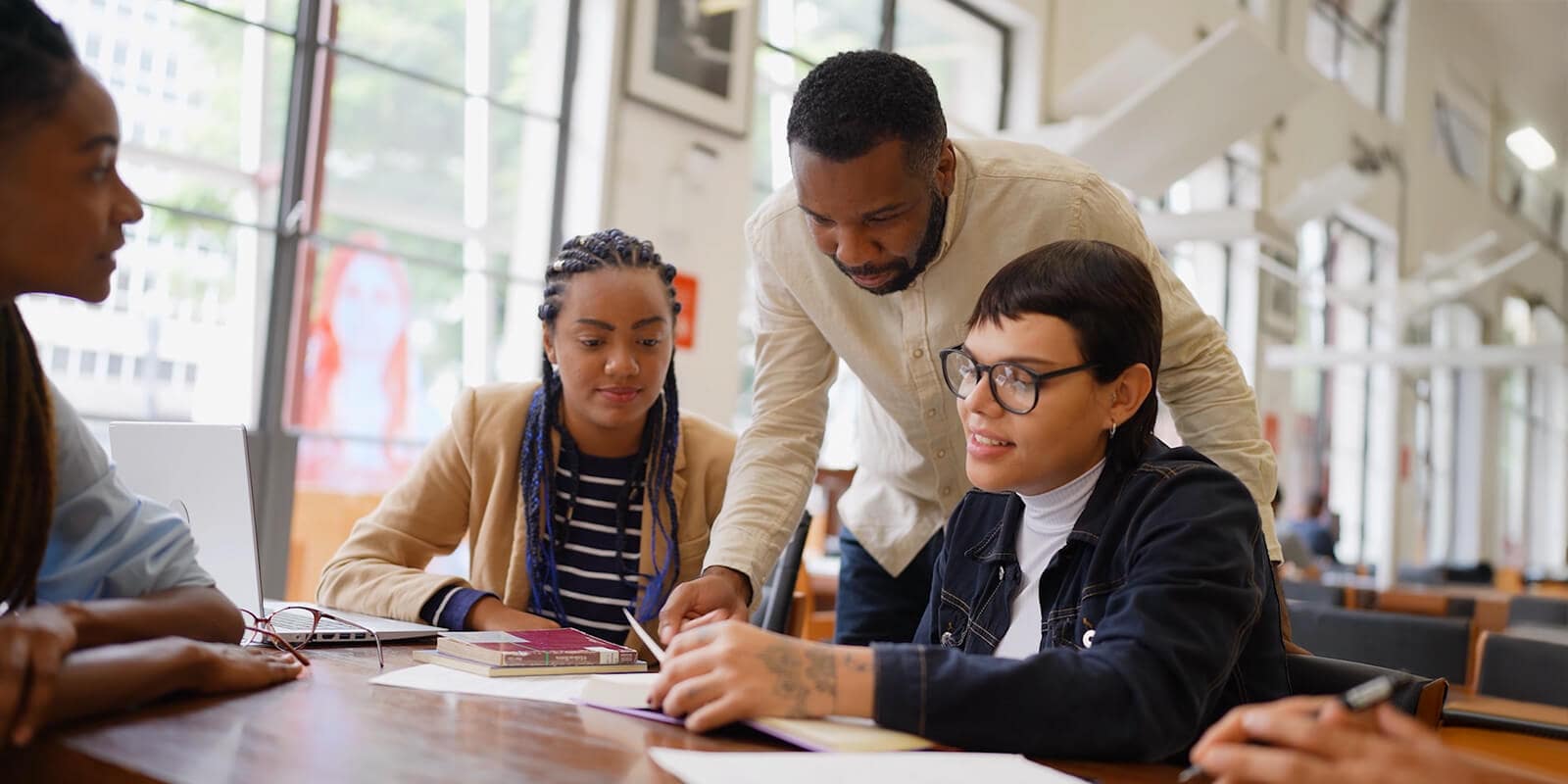 A diverse group of students collaborates around a table with books and laptops in a bright, modern library setting.