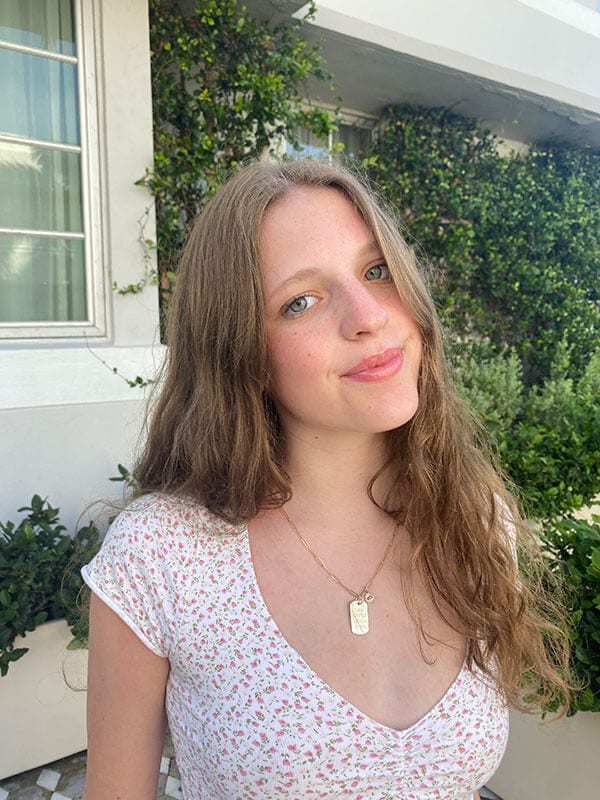 Blog author Cassidy is standing outside in front of an ivy-covered wall. She has long light brown hair and is wearing a short-sleeved white shirt with a red floral print.