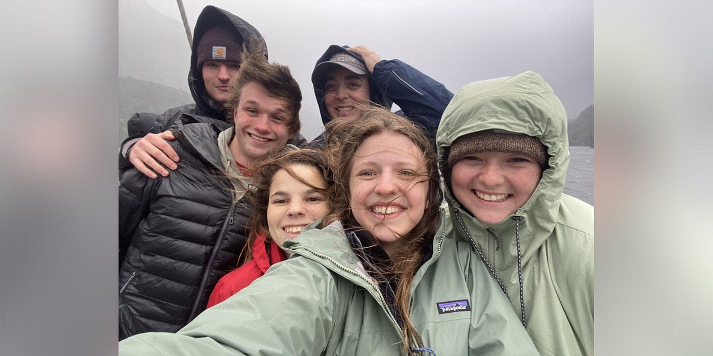 A group of six college students wearing coats and hats stand outside in front of a lake on a windy day.