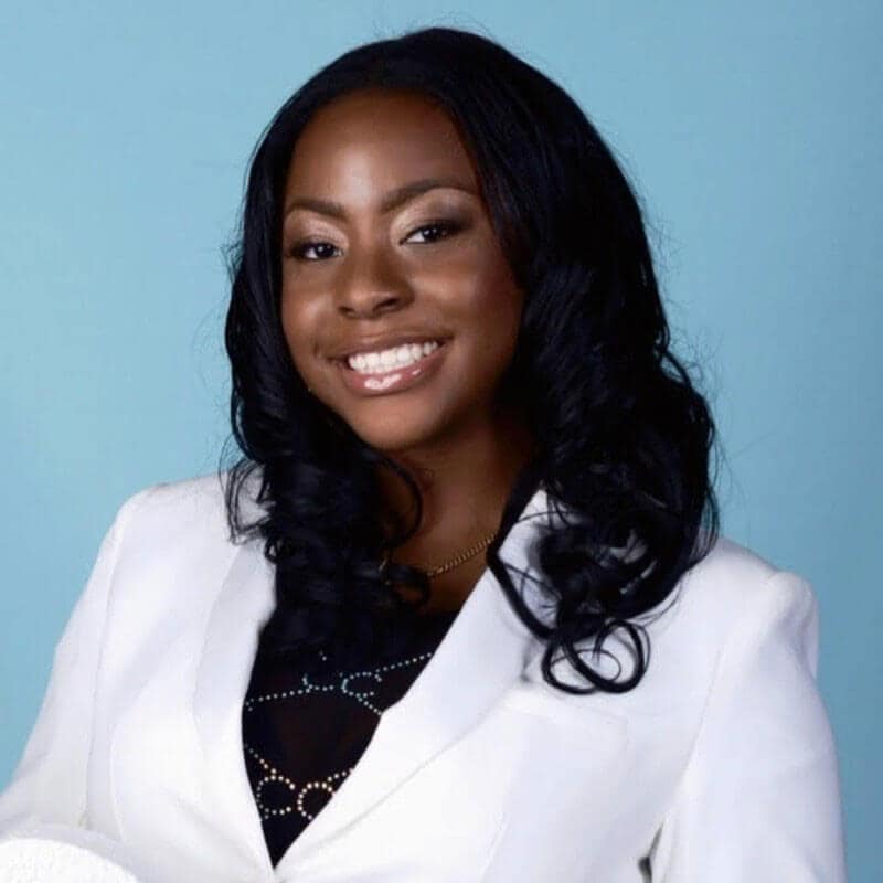 Blog author Cydell is smiling in front of a blue background. She has shoulder-length dark wavy hair and is wearing a white blazer over a black shirt.
