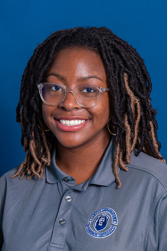 A studio headshot of blog author Cydell. She has medium-length braids and is wearing clear framed glasses and a grey polo shirt.