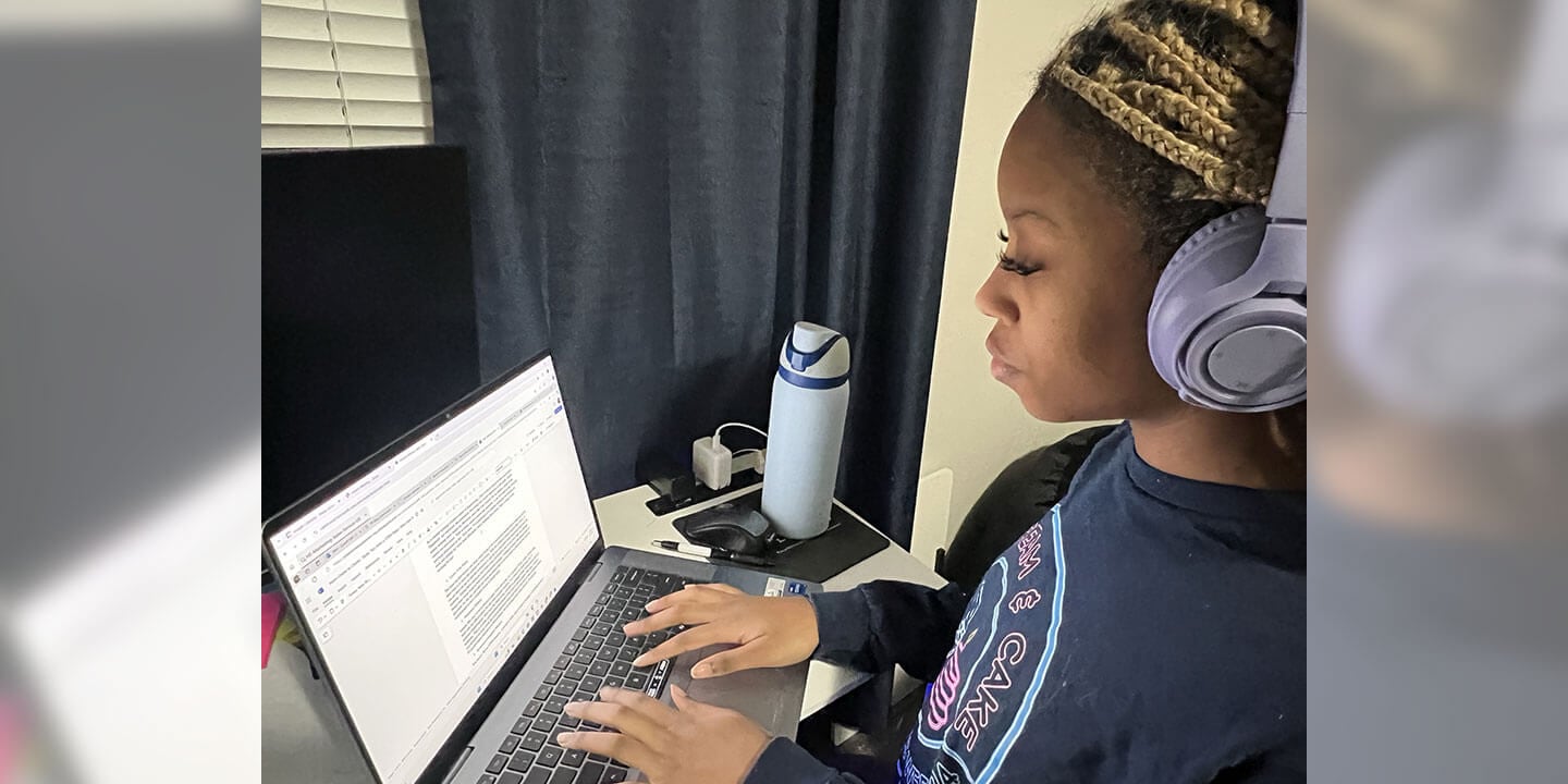 A student wearing noise-cancelling headphones sits at a desk working on a laptop.