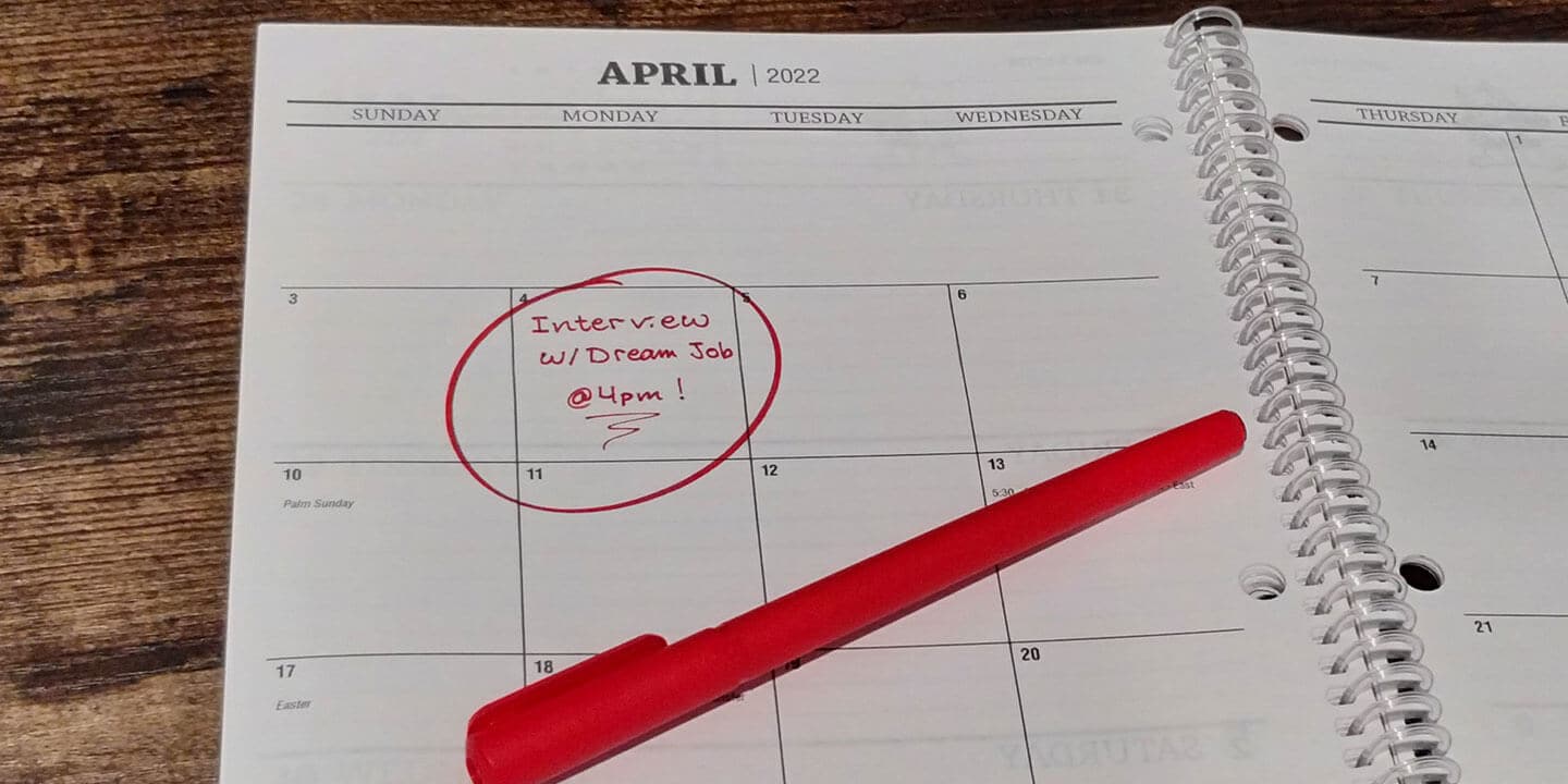 An open planner on a desk with the words ‘Interview with dream job @ 4 pm!’ written in red ink on one of the days. 
