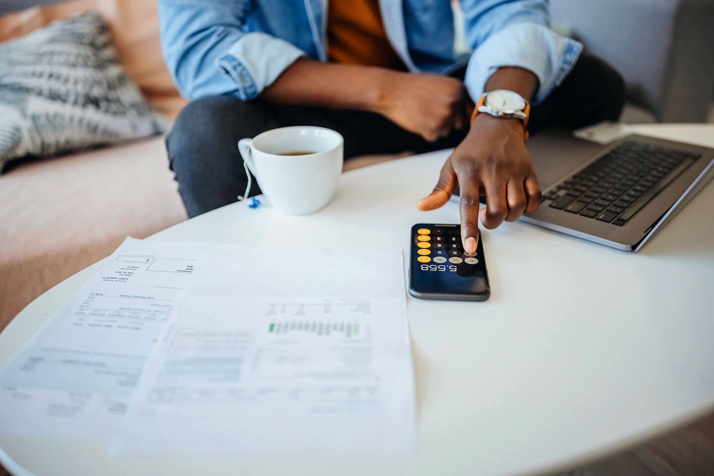 An adult with financial papers and a computer sitting on the table in front of them uses the calculator on their phone. 