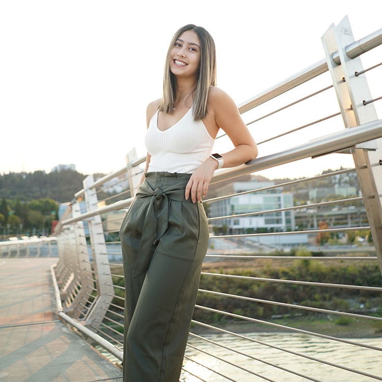 Blog author Grace Oh is smiling. She has long brown hair, white tank top, green pants and leaning on a fence. 