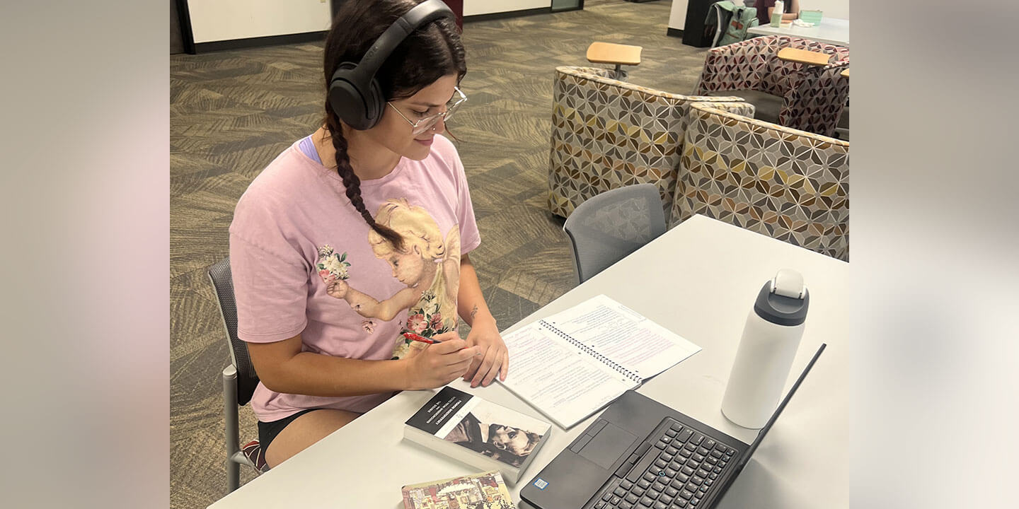 A young woman wearing headphones sits at a desk with a book, an open laptop, a composition notebook, and a water bottle.