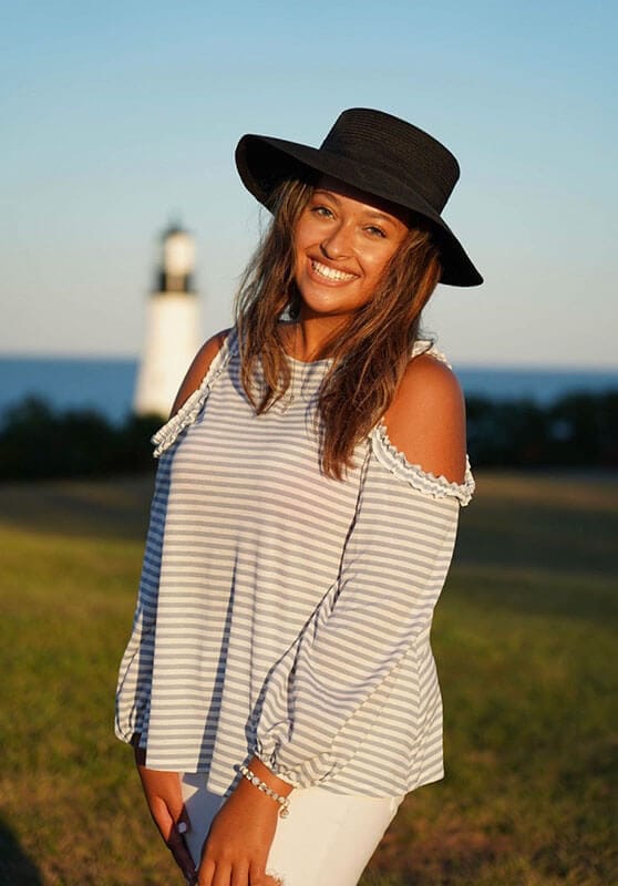 Blog author Jasmine is standing outside with a lighthouse in the background. She is wearing a fedora-type hat and a striped long sleeve top.