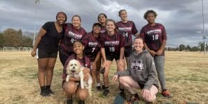 A group of university students from an ultimate frisbee team are standing arm-in-arm outside. Another team member is crouching down in front of the group with a small dog on her lap. 