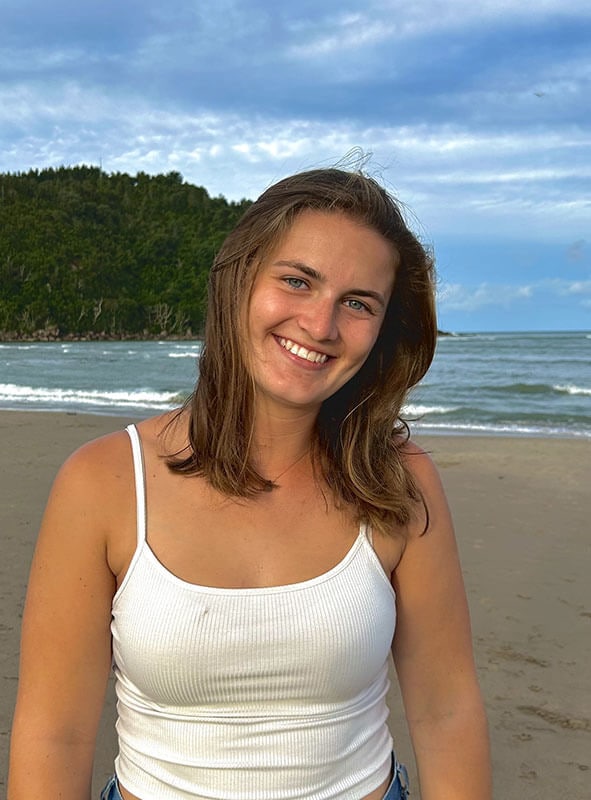 Blog author Kennedy is standing outside on a beach. She has shoulder-length dark blonde hair and is wearing a white tank top.