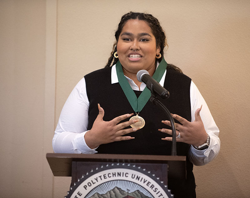 Blog author Katie is speaking from a podium. She is wearing a black vest over a white blouse and has a green medal around her neck.
