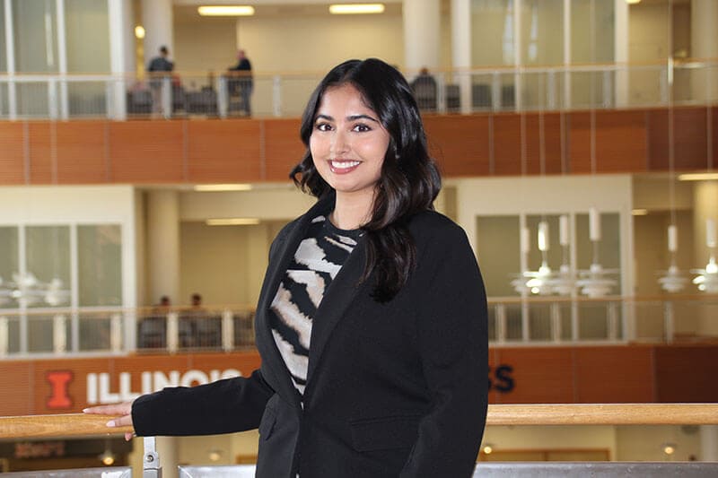Blog author Keya is standing by a railing in a campus building. She has long dark wavy hair and is wearing a black blazer over a white and black patterned blouse.