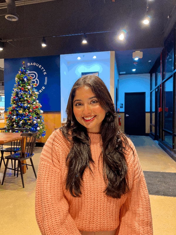 Blog author Keya is standing in a food court area on her campus. There is a Christmas tree in the corner. Keya has long dark hair and is wearing a peach-colored sweater.