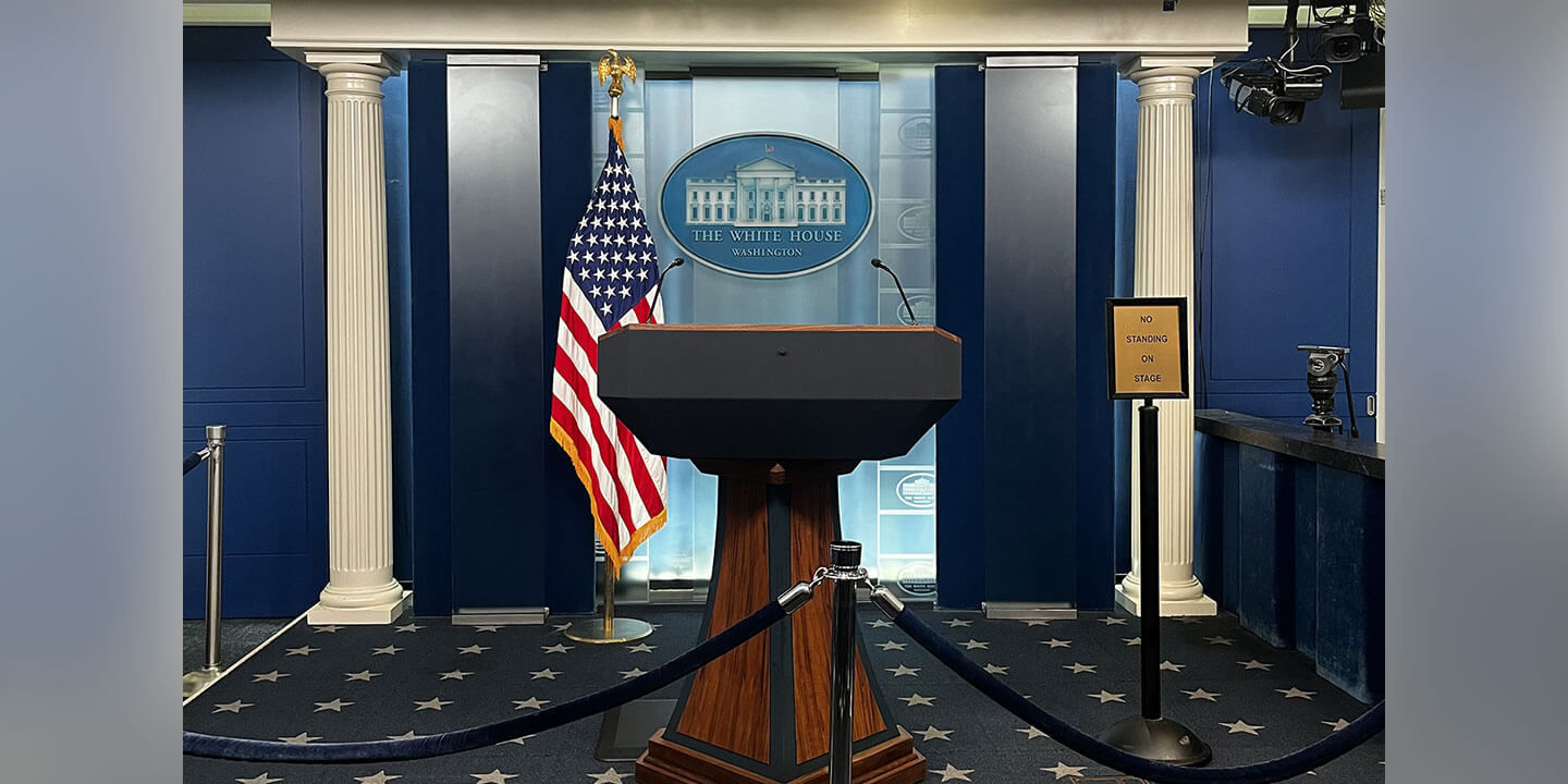 A podium behind a blue velvet rope barrier. An American flag is posted behind on the left and signage reads “The White House Washington”.