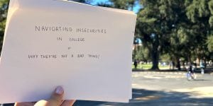 A person’s hand holding up a white piece of paper with the words: “Navigating Insecurities in College + Why They’re Not a Bad Thing”.