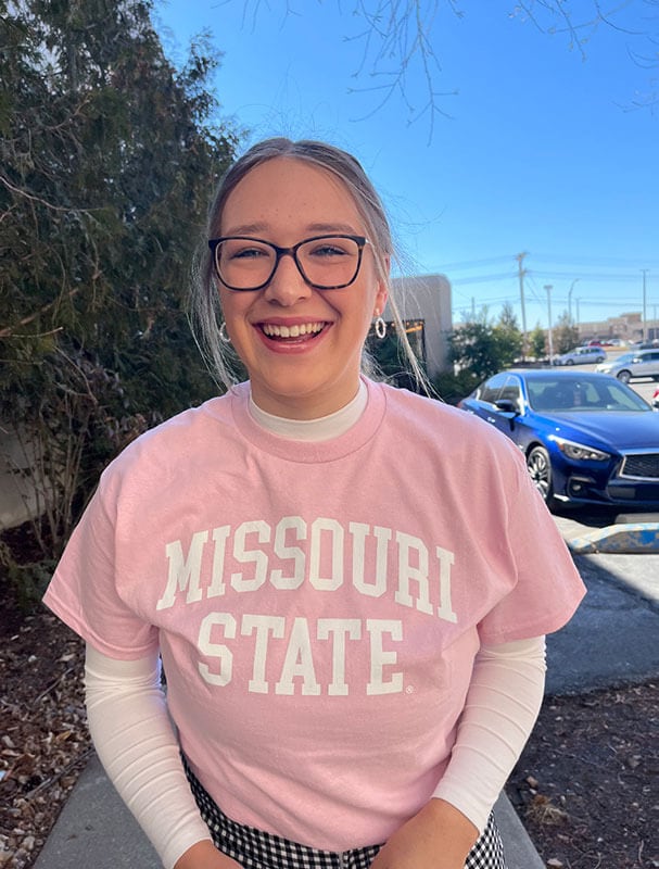 Blog author Molly is standing outside wearing a pink t-shirt with white lettering spelling ‘Missouri State’. She has blonde hair pulled back and is wearing dark framed eyeglasses.