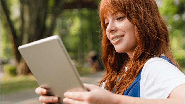 A woman sitting outdoors is smiling at a tablet device screen.