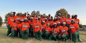 Members of Florida A&M’s marching band gather for a group photo on a football field. They are wearing matching baseball caps, orange t-shirts and green pants.