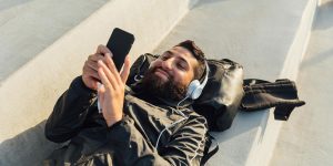 A young man lies on his back outside on a concrete step. He is wearing headphones and using an app on his phone.