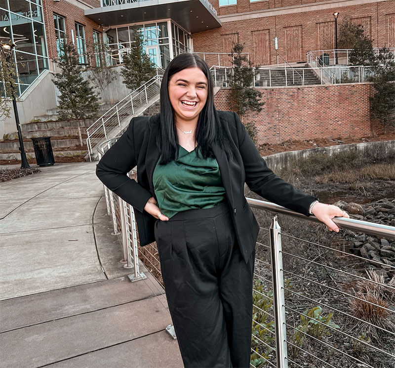 Blog author Rinn is smiling and leaning against a walkway railing. She has long brown hair and is wearing a black pantsuit over a green top.