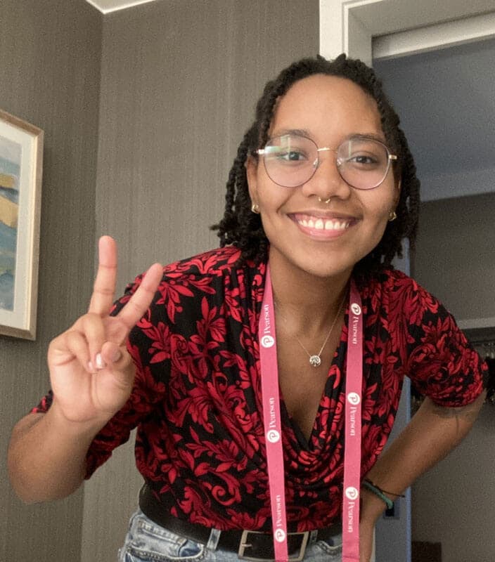 Blog author Rachel is smiling and holding up her right hand in a ‘peace sign’. She has short dark braided hair and is wearing a blue lanyard on her neck.