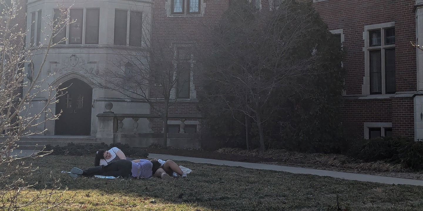 A small group of college students lie in the grass in front of an academic building.