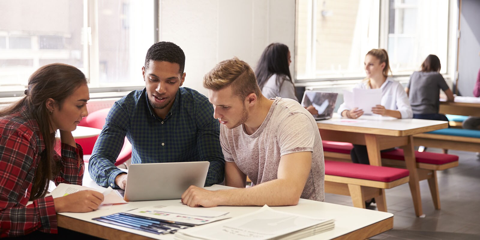 Three students sit at a table in a college common room. They are gathered around a laptop computer and are looking at the screen with interest.
