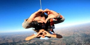 Two people skydiving in tandem. They are both making a heart shape with their hands.