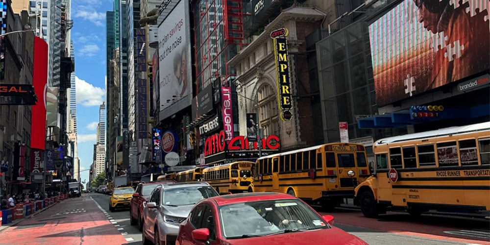A street view in New York City with tall buildings, lighted signs, and cars and busses in the street.
