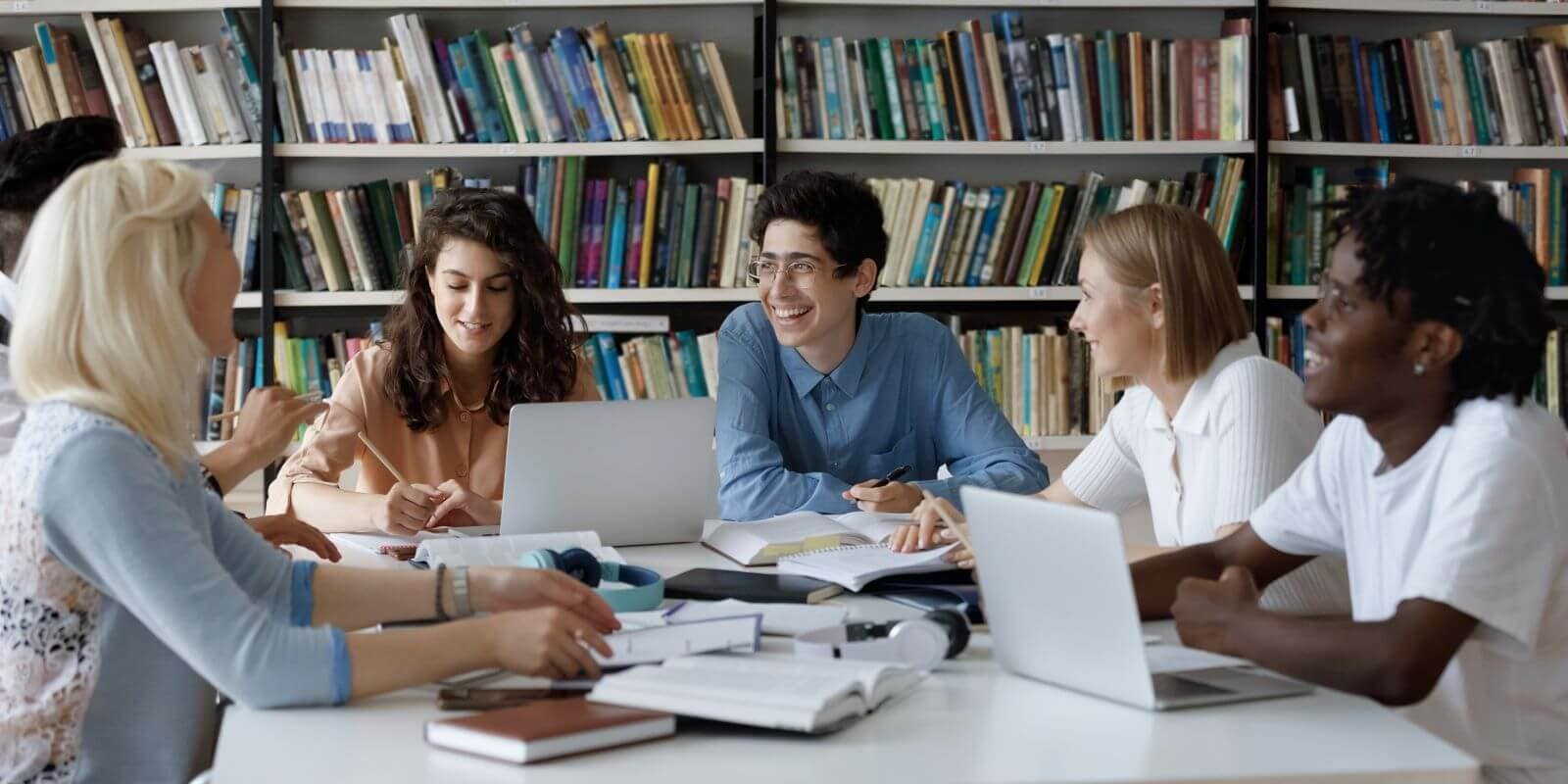 A diverse group of college-aged students sits around a table full of books and laptops in a library