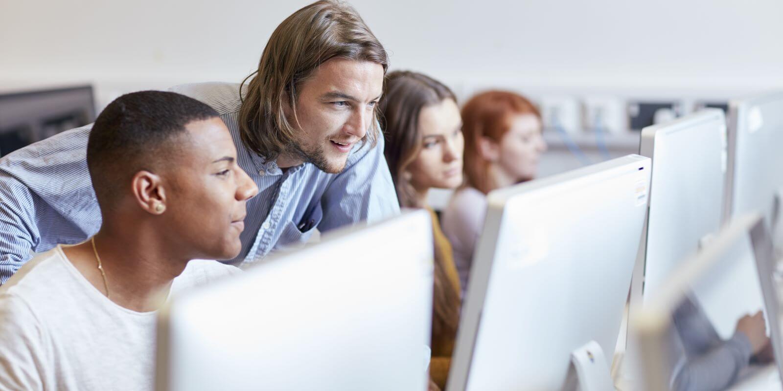 A row of computers in a computer lab with studentsw and instructor looking at content