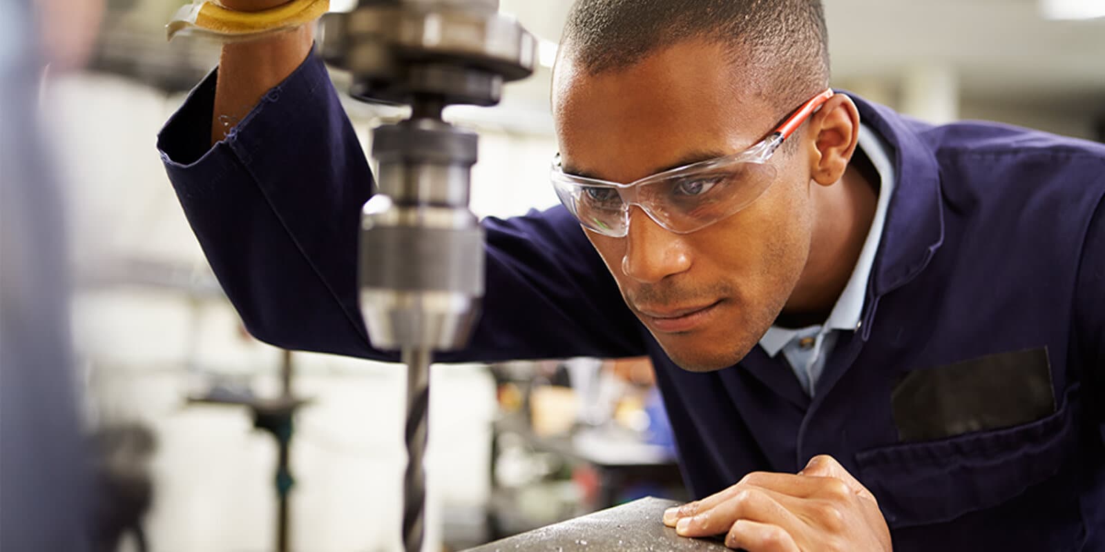A teacher in a suit interacts with three students at a classroom table, holding books and pens, with a bright window in the background.