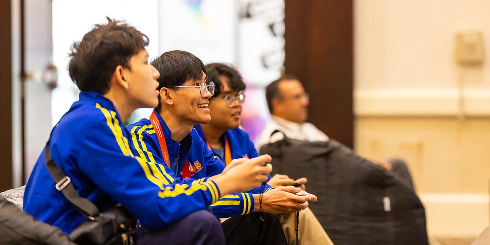 Three high school students in a well lit room, interacting with each other while playing videogames.