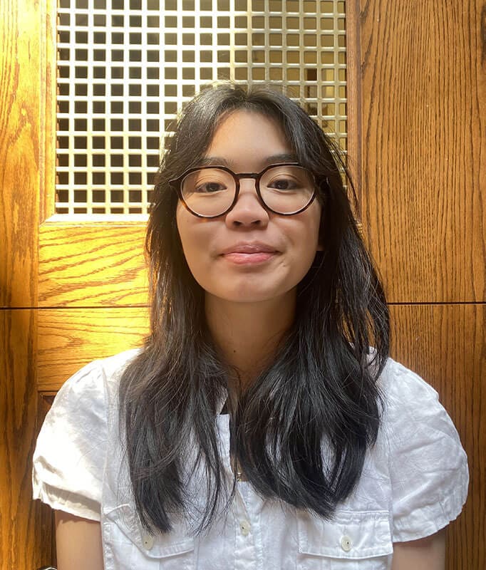 The blog author, Alexis, has long dark hair, is wearing glasses, and a white blouse. She is standing in front of a wooden wall with a grid pattern.