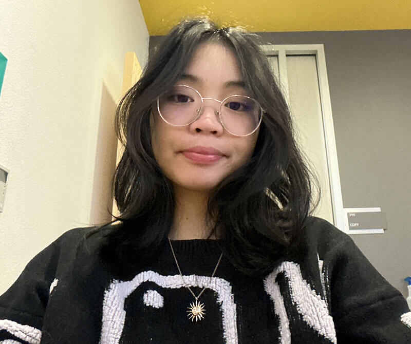 The blog author has long dark hair and is wearing glasses, a sweater with a white graphic pattern, and a gold sun necklace.