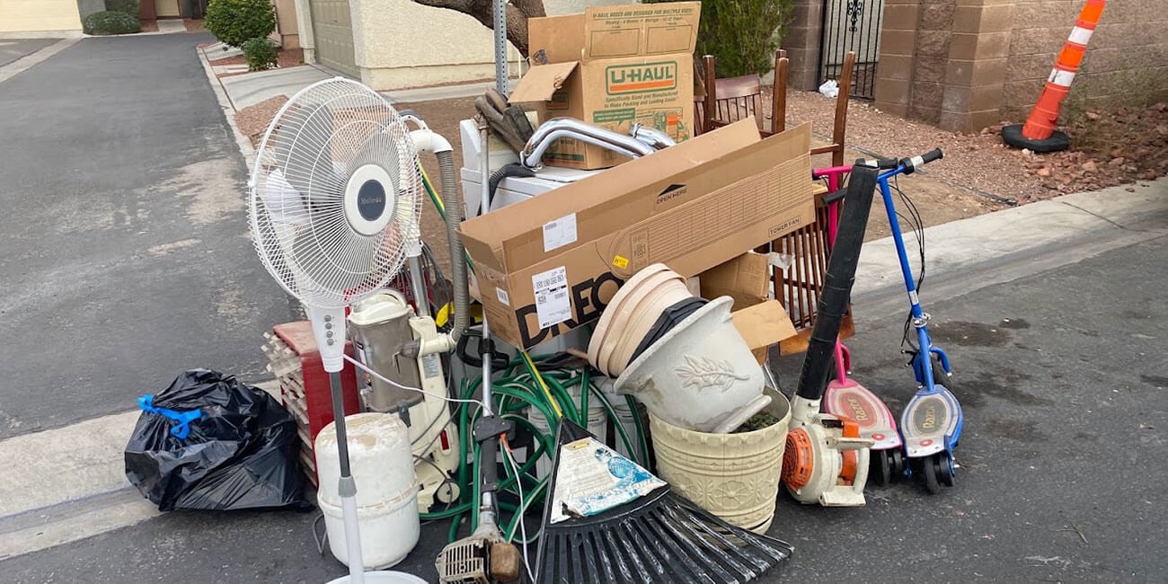 Image shows a pile of items left at a curb likely trash day for garbage pickup including boxes, a standing, a rake, a leaf blower, two toy scooters, and a trash bag among other items.