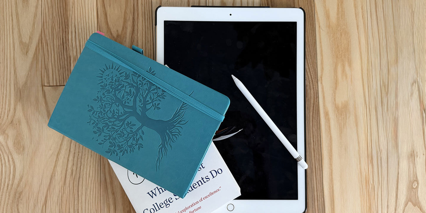 An iPad, stylus, journal, and a book positioned on a wooden surface.