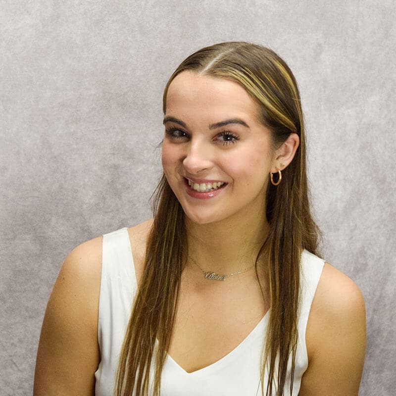 The blog author has long light brown hair, is wearing a sleeveless white dress, a gold hoop earring, and a necklace with the name “Alaina” on it.